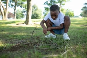 A black boy in jeans and a t-shirt crouched down poking at the ground with a stick.