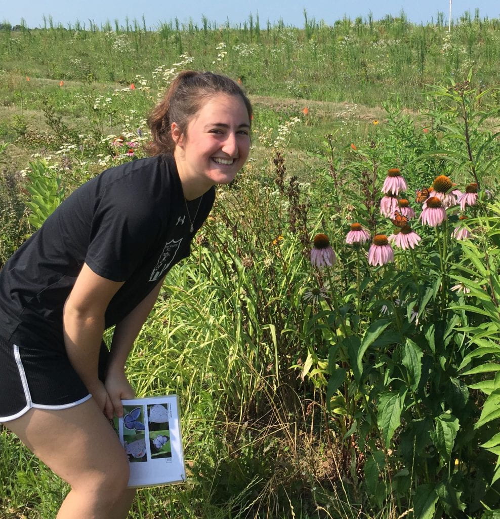 Smiling young woman holding a sheet with pictures of butterflies, and standing next to blooming purple coneflowers. You can see a field in the background.