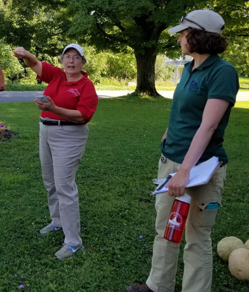 Woman on left is wearing a red shirt, talking, and gesturing with her hands. Woman on right in wearing a green shirt and watching and listening to the woman on the left.