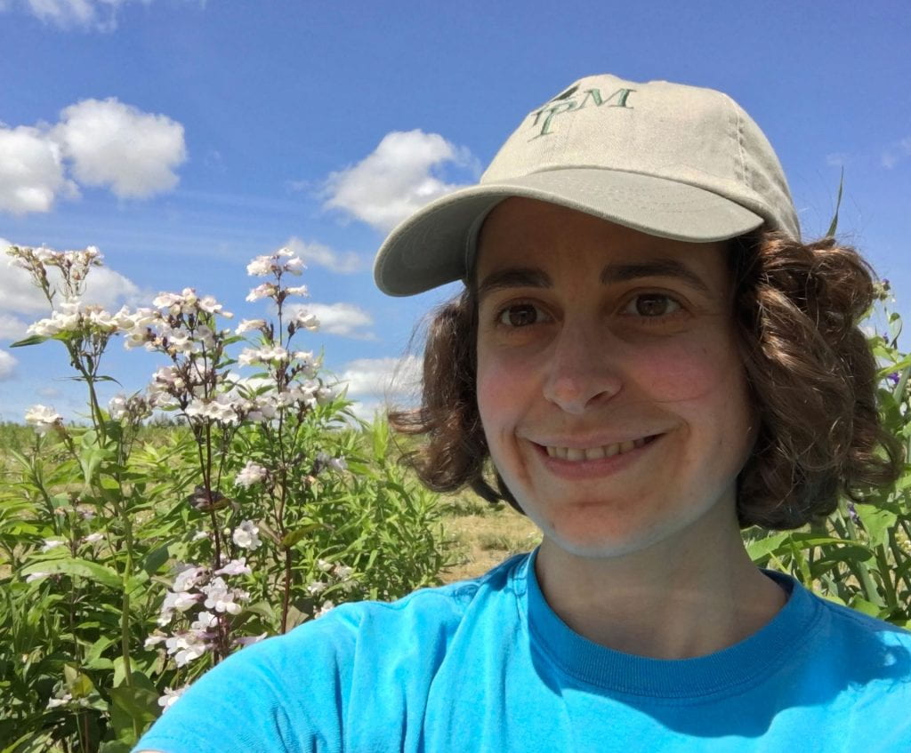 Woman in a blue shirt and baseball cap looking into the camera. In the background you can see white bell-shaped flowers and blue sky with a few puffy clouds.