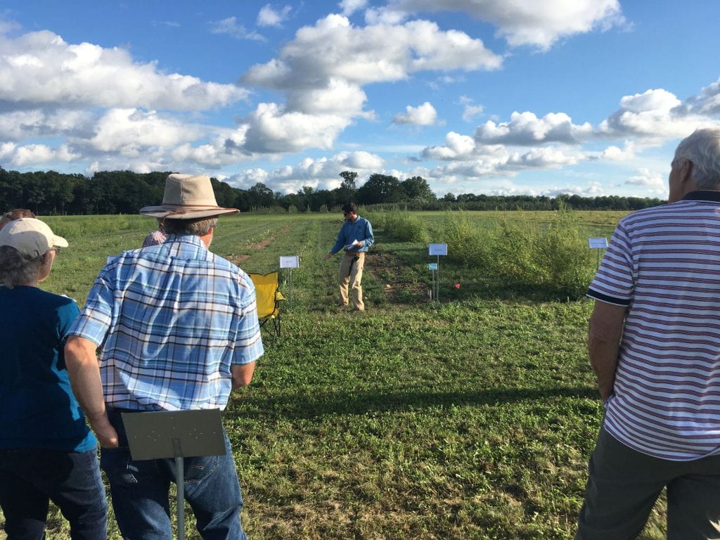 Researcher points to labeled rows of Christmas trees growing in a field, while meeting attendees watch