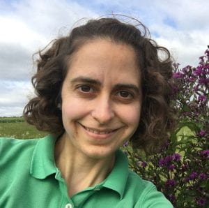 head shot of a white woman with brown curly hair in a green polo shirt; taken in a field with deep magenta flowers in the background
