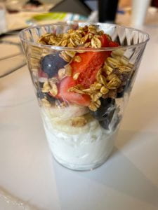 A clear plastic cup with a layer of yogurt on the bottom topped with sliced strawberries, bananas, apple, blueberries, and granola.