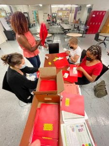 A birds-eye-view photo of a table with folders, stickers, and handouts with Miriam, Sashana, Jackie, and Jaylyn working to fill the folders.