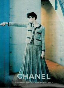 Print ad from Chanel featuring a model wearing a jacket and long, pleated skirt. Both jacket and skirt are teal in color; the jacket has black trim along edges and the four pockets. The model is facing center with head turned to the left and left arm extended to the side. 