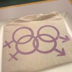 This image is of a silk screened t-shirt with two male and two female signs interlinked; the symbols are lavender in color.