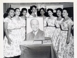 Eight young female Hispanic or Latina campaign workers posing with a photo of President Eisenhower. They are all wearing dresses with the word "IKE" written all over. 
