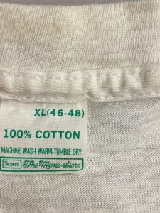 Clothing tag from a t-shirt. Reads "100% cotton, machine wash warm tumble dry, SEARS, The Men's Store." 