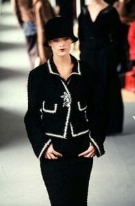 A model walking down the runway wearing a 1998 Chanel jacket, black skirt, and black hat. The jacket is black with white trim around the edges and pocket. The center of the jacket is held together with a grey, flower brooch.