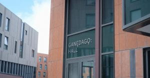 Photo of a modern building made of glass and light brown panels with the words Ganedago Hall above a set of doors.