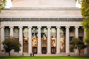 Image of MIT's Killian Court which is a large pillared building with floor to ceiling glass windows in between the pillars. The building also has a large dome on top.