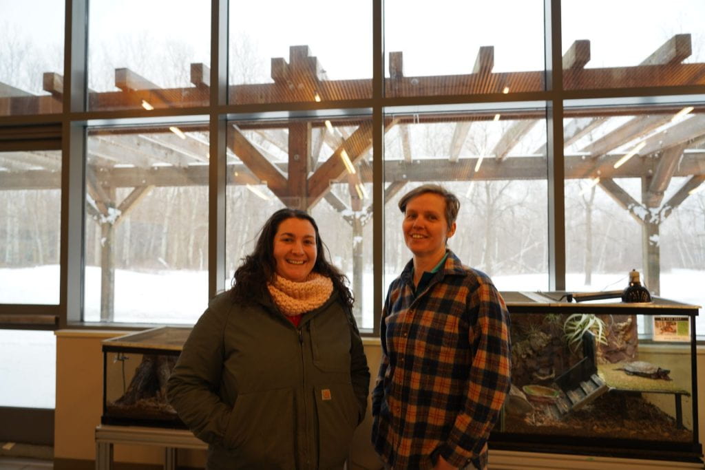 Two women standing in front of a window and nature exhibit. 