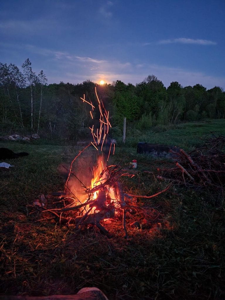 An outdoor fire in a fire pit, with the moon rising over a background of pasture and forest.