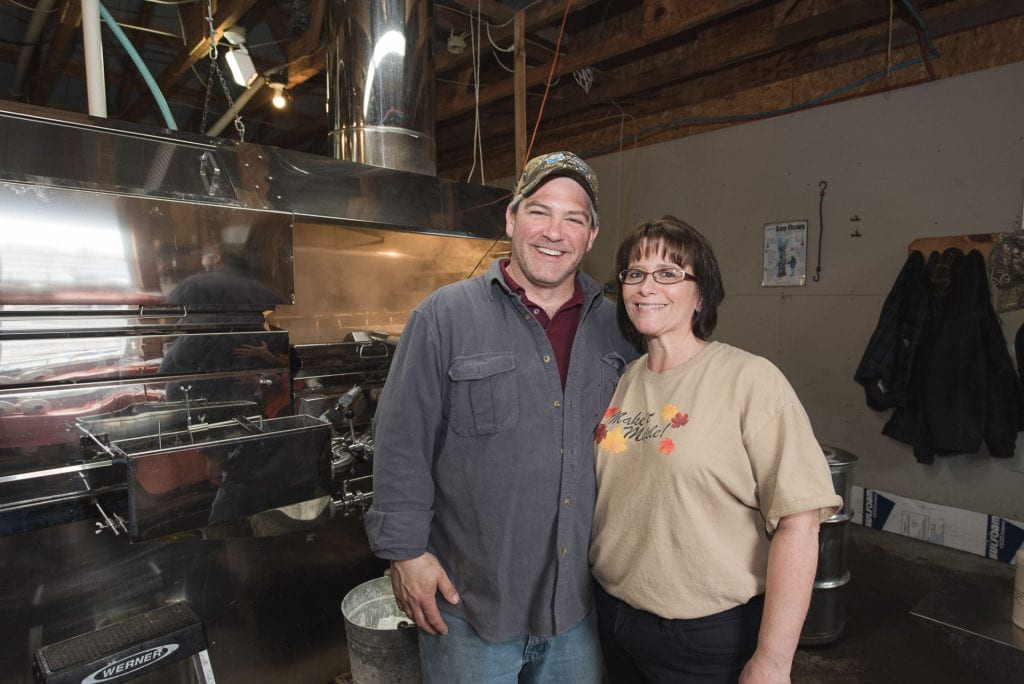 Photo of a man and woman standing in front of a maple syrup evaporator. 