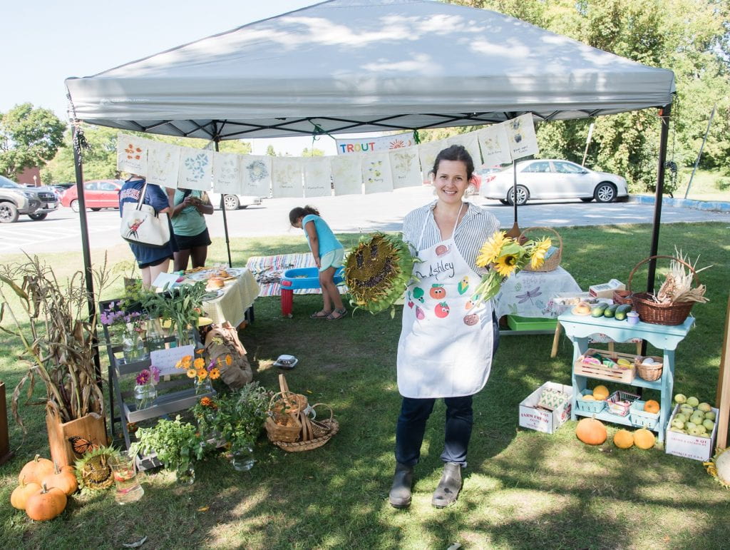 Photo of a woman standing at her market tent with assorted herbs, vegetables, holding sunflowers.