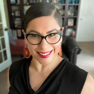 Sara in glasses, red earrings and a black shirt in front of a bookcase