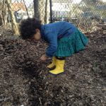 A small child in a jean shirt, teal skirt and bright yellow rain boots put seeds in the ground