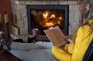 Photo: Person with long brown hair, wearing comfy socks and sweat pants, sitting on the floor in front of a blazing fire place reading a book as a black and white cat rests its head on their leg