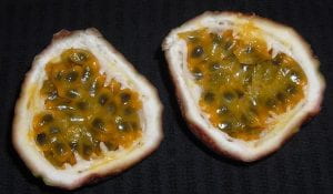 Photo: Cross sections of a passion fruit. The fruit has been sliced in half. There is a thick white skin and the inside is full little black seeds with a gelatinous yellow coat.
