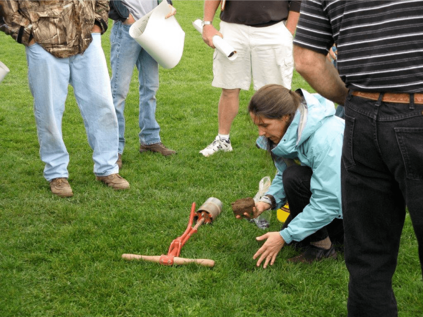 Photo shows Jen Grant kneeling to explain the use of a cup cutter to scout for grubs on turf as others watch