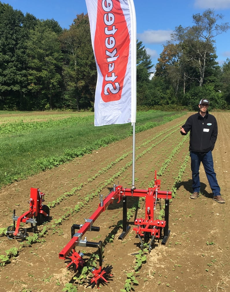 Image shows a sales rep from Steketee company, standing next to a pull behind cultivator that uses a multi-faceted method of disturbing soil and uprooting weeds