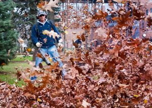 image of a man in long pants and sleeves, a baseball cap, and ear coverings using a leaf blower. In front of himn leaves are being blown towards the camera.