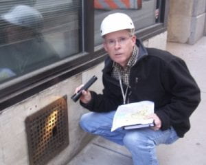 photo of Bobby Corrigan wearing a hard hat, holding a clipboard in one hand and a flashlight in the other pointing out a rusted wall grid plate with a hole large enough for a rat to fit through.
